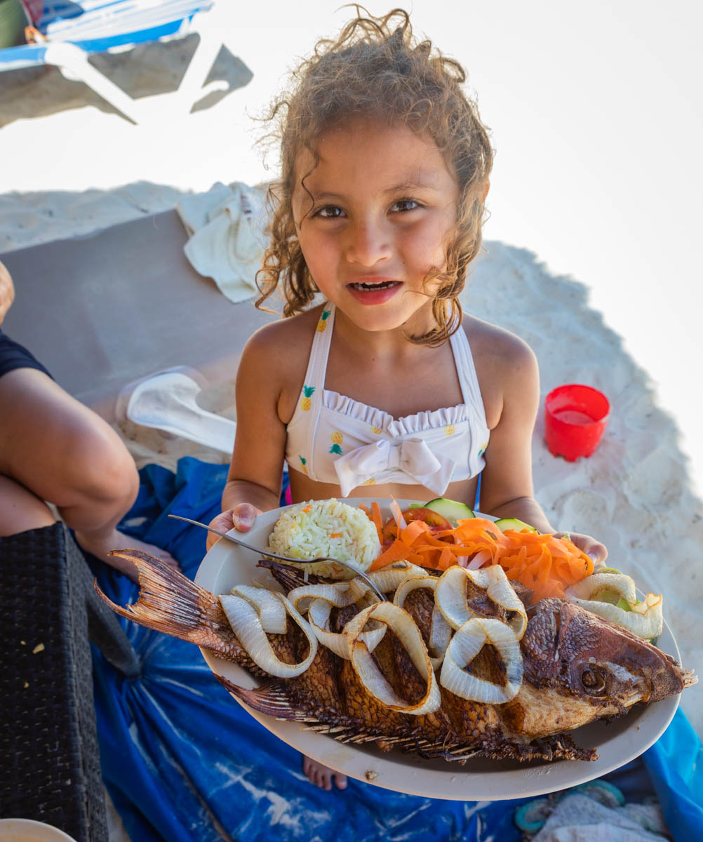 A child holds a plate overflowing with a whole fried fish.